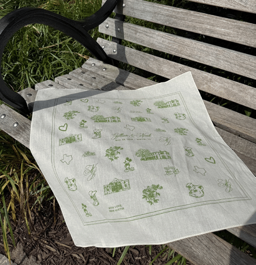 A light-colored square cloth with green illustrations of buildings, hearts, Mickey Mouse, Texas, flowers, and initials J & N is laid on a wooden park bench.