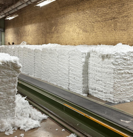 Large stacks of compressed white cotton bales are lined up along a green conveyor belt in a factory with brick walls and overhead lighting. Loose cotton pieces are scattered on the floor.