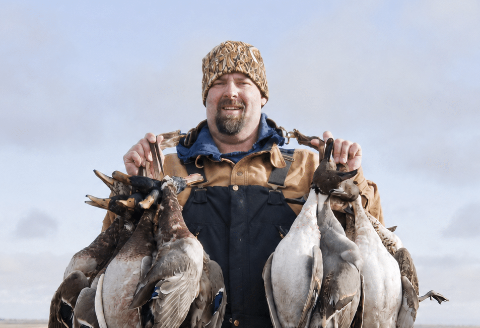 A man in a brown jacket and patterned beanie smiles while holding several ducks by their necks in each hand, standing outdoors under a cloudy sky. A man in a brown jacket and patterned beanie smiles while holding several ducks by their necks in each hand, standing outdoors under a cloudy sky.