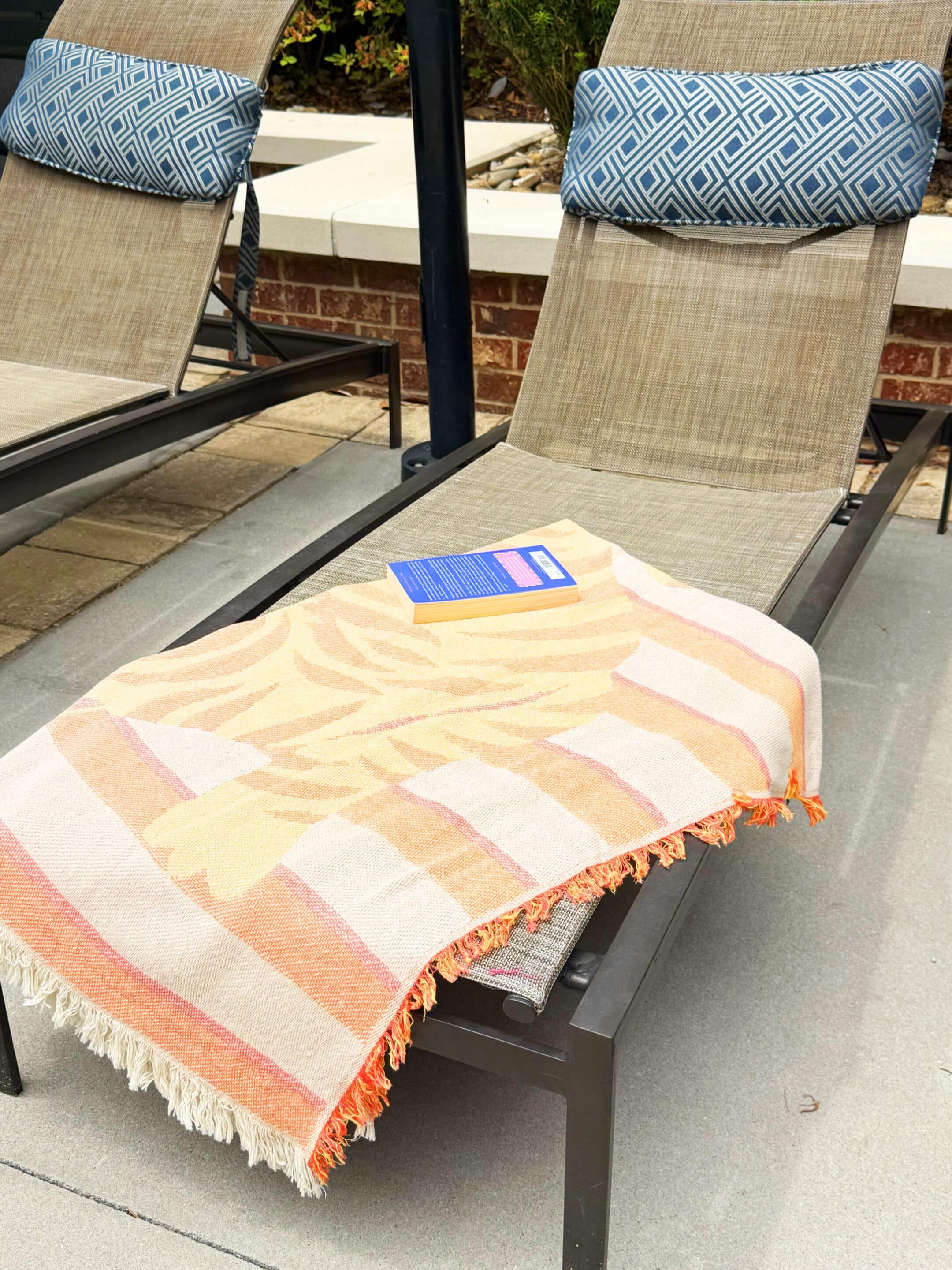 Two outdoor lounge chairs with beige fabric and blue geometric pillows, a peach-and-pink striped blanket, and a small blue book resting on the chair.