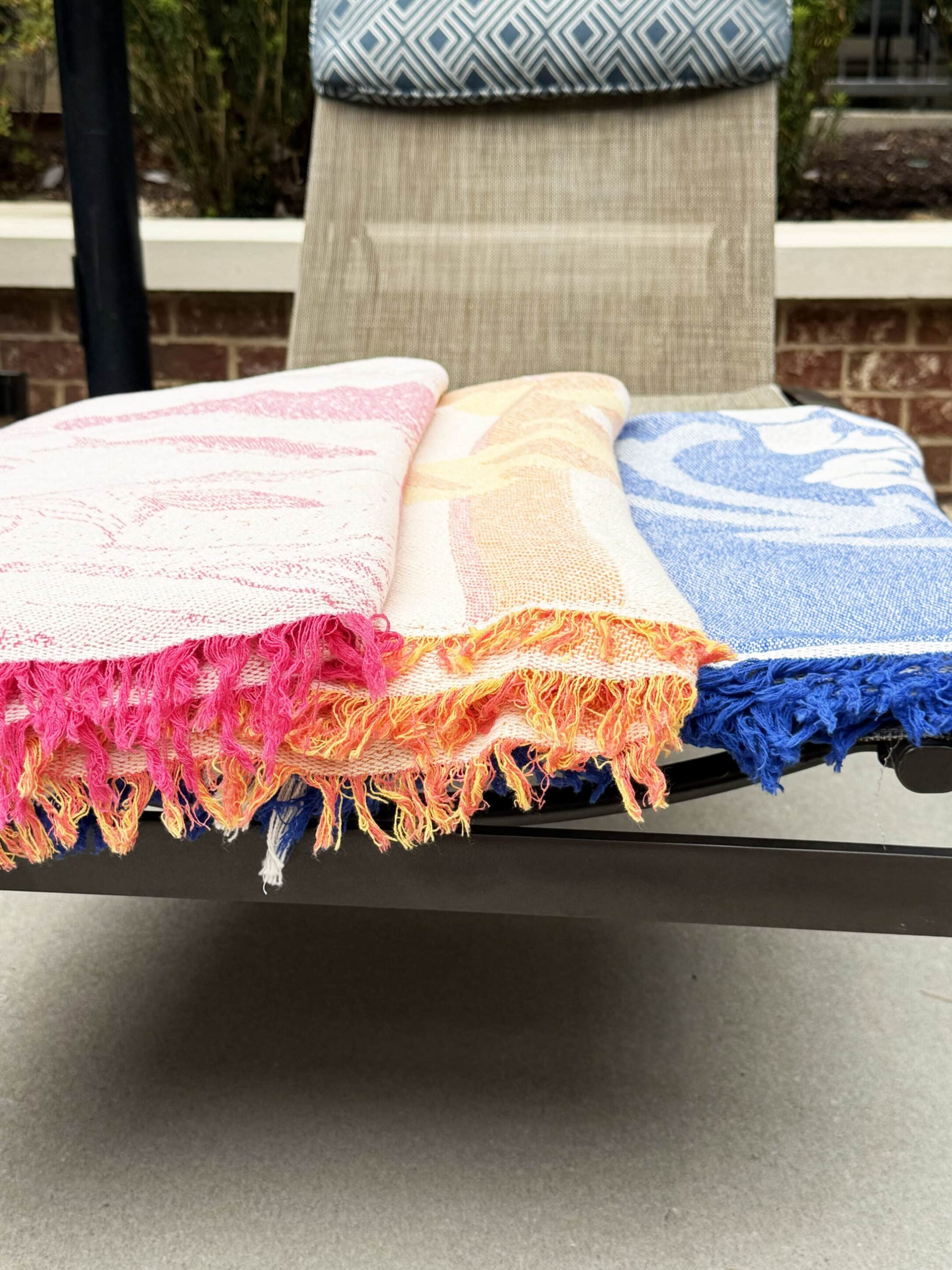 Colorful beach towels draped over a lounge chair outdoors, pink/orange/blue fringe hanging over the edge.