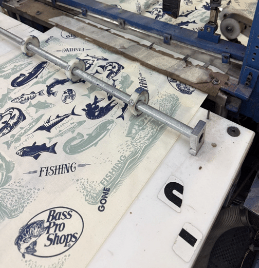 A close-up of a printing press producing sheets with Bass Pro Shops, fish illustrations, and the word FISHING in blue ink on cream-colored paper or fabric. A metal rod with bolts holds the sheets in place.