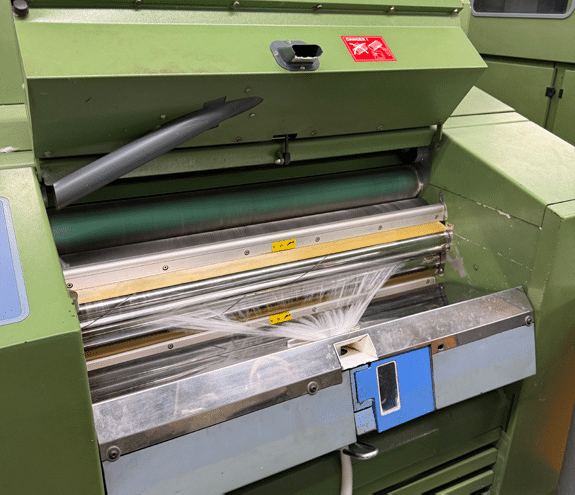 A close-up of a green industrial machine with spinning rollers, tangled white plastic film caught between them, and control buttons visible on the front panel. A warning sticker is displayed above the rollers.