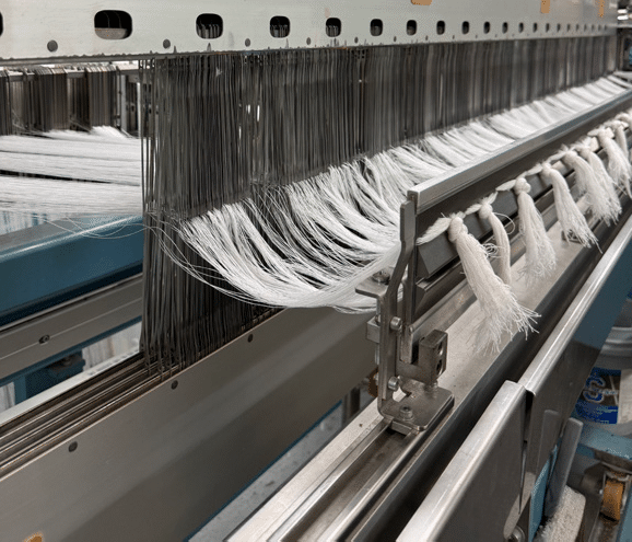 Close-up of a textile weaving machine with white threads passing through metal heddles and guides, showing the process of weaving fabric in an industrial setting.
