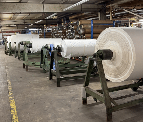 Large rolls of white paper are lined up on metal stands inside an industrial warehouse, with shelves of pipes and other equipment visible in the background.