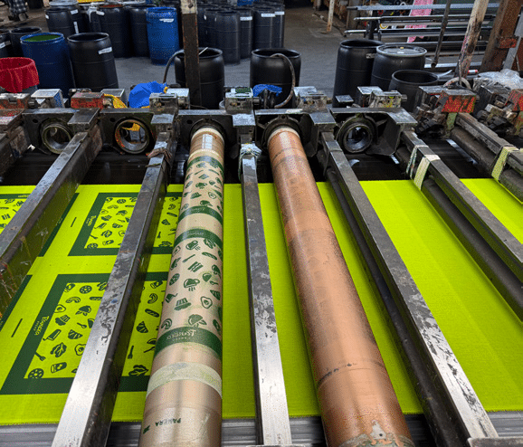 A close-up of a textile printing machine with cylindrical rollers printing green fabric with various black icons in a factory setting. Black barrels and other equipment are visible in the background.